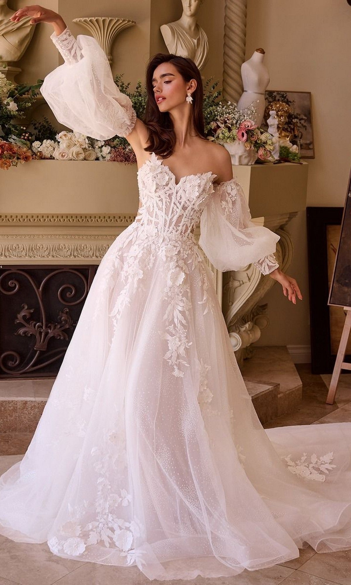 A woman poses indoors before floral decorations, wearing the Andrea and Leo WL018 long wedding dress featuring a white embroidered design, sweetheart neckline, off-shoulder style, and sheer puffy sleeves.